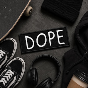 May include: A rectangular black embroidered patch with the word DOPE stitched in white thread. The patch is surrounded by a black skateboard, black sneakers with white laces, a black beanie, black headphones, and a black hooded sweatshirt.