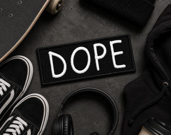 May include: A rectangular black embroidered patch with the word DOPE stitched in white thread. The patch is surrounded by a black skateboard, black trainers with white laces, a black beanie, black headphones, and a black hooded sweatshirt.