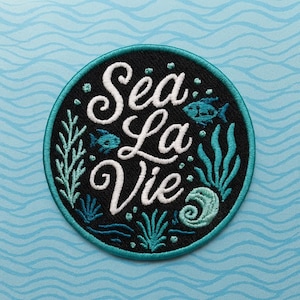 Op de afbeelding: Ronde geborduurde patch met de woorden "Sea La Vie" in witte letters op een zwarte achtergrond. De patch heeft turquoise accenten, waaronder vissen, zeewier en een rand, tegen een lichtblauwe golvende achtergrond.