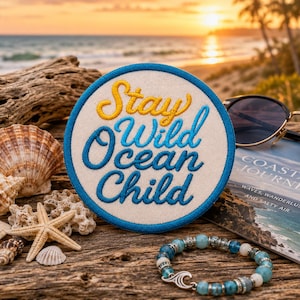 Peut inclure: Un écusson brodé circulaire avec un fond crème et une bordure bleue. Le texte "Stay Wild Ocean Child" est cousu en lettres cursives jaunes et bleues. L'écusson est placé parmi des coquillages, une étoile de mer, un bracelet et un livre.