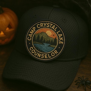 May include: A navy blue baseball cap with a circular embroidered patch. The patch features a landscape scene with the text "CAMP CRYSTAL LAKE COUNSELOR". A carved pumpkin and a plastic spider are in the background.