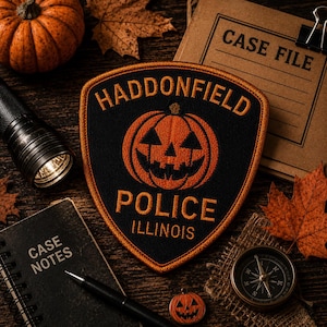 May include: A shield-shaped embroidered patch featuring a black background with an orange border. The text reads HADDONFIELD POLICE ILLINOIS in orange, centred above and below a large orange jack-o'-lantern graphic with black facial features.