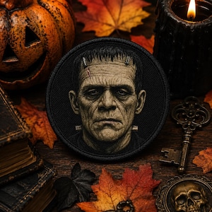 May include: A circular embroidered patch featuring a detailed portrait of Frankenstein's monster in shades of grey and green. The design includes the character's signature flat head, heavy brow, facial scars, and metallic neck bolts on a black background.