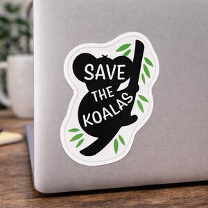May include: A white sticker on a laptop with a black silhouette of a koala bear holding onto a branch. The sticker has the words "SAVE THE KOALAS" in white text. Green leaves are around the koala and branch.