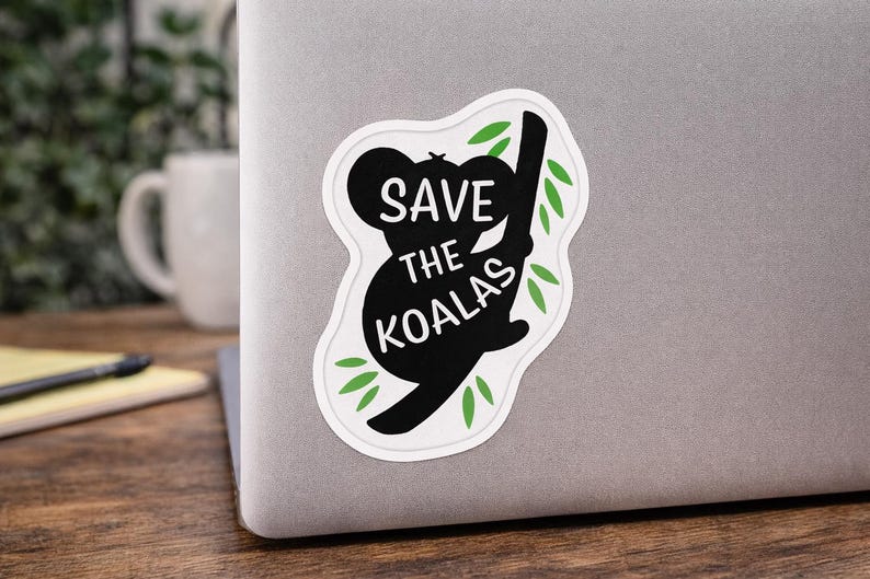 May include: A white sticker on a laptop with a black silhouette of a koala bear holding onto a branch. The sticker has the words "SAVE THE KOALAS" in white text. Green leaves are around the koala and branch.