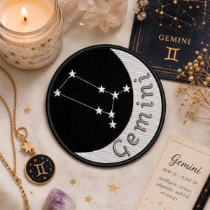 Puede incluir: Parche redondo negro y blanco con la constelación de Géminis en blanco, sobre un diseño de media luna. La palabra "Gemini" está bordada en gris. También se ven una vela y una tarjeta del zodiaco.