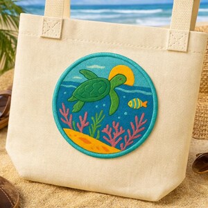 以下が含まれることがあります： クリーム色のトートバッグ。円形の刺繍パッチには、青い海の中で泳ぐ緑色のウミガメが描かれています。パッチには黄色い太陽、小さな黄色い魚、サンゴが含まれています。バッグにはクリーム色のハンドルが2つ付いています。