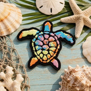 May include: Embroidered sea turtle patch with a black border, featuring a colourful design of blue, pink, yellow, and orange. The patch is surrounded by seashells, a starfish, and a fishing net on a light blue wooden surface.
