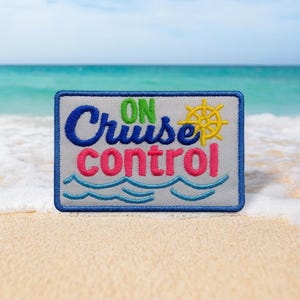 May include: Embroidered patch with the words "ON Cruise control" in blue, green, and pink, with a yellow ship's wheel. The patch has a blue border and wavy lines, set against a beach and ocean backdrop.