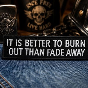 May include: A black embroidered patch with white text reading "IT IS BETTER TO BURN OUT THAN FADE AWAY". The patch is displayed on denim, with a skull lighter and other biker-themed items visible.