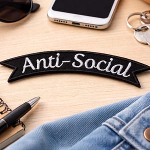 Puede incluir: Un parche negro bordado con las palabras "Anti-Social" en blanco. El parche está sobre una superficie de madera clara, junto con gafas de sol, un teléfono inteligente, llaves, un cuaderno, un bolígrafo y tela vaquera.