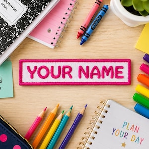 May include: A rectangular patch with the text "YOUR NAME" in pink, surrounded by a white border and a pink frame. The image also includes school supplies like notebooks, crayons, and colored pencils, and sticky notes with positive messages.