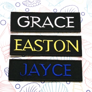 Puede incluir: Tres parches de tela negra con letras bordadas en blanco, amarillo y azul. Los parches dicen "GRACE", "EASTON" y "JAYCE".