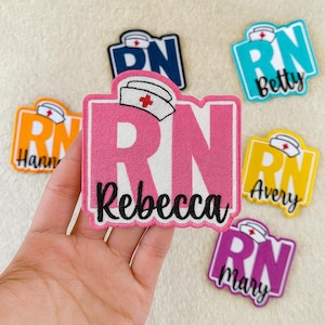 May include: Embroidered RN name badges in a variety of colours. The central badge is pink with "Rebecca" in black script. Other badges include "Betty," "Avery," "Mary," and "Hann." Each badge features a nurse's cap and the letters "RN."