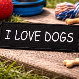 May include: A black rectangular patch embroidered with the words "I LOVE DOGS" in white. The patch is on a wooden surface, with dog treats, a red ball, a blue bowl, and a rope toy in the background. Green grass surrounds the objects.