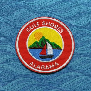 May include: A round embroidered patch with a red border and the words "GULF SHORES ALABAMA". The patch features a sailboat on blue water, green mountains, and a yellow sun.