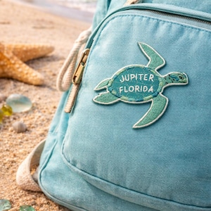Pu&ograve; includere: Zaino azzurro con una toppa a forma di tartaruga con la scritta "Jupiter Florida". Lo zaino ha una tasca frontale con cerniera dorata e spallacci beige. Lo sfondo mostra una spiaggia sabbiosa e l'oceano.