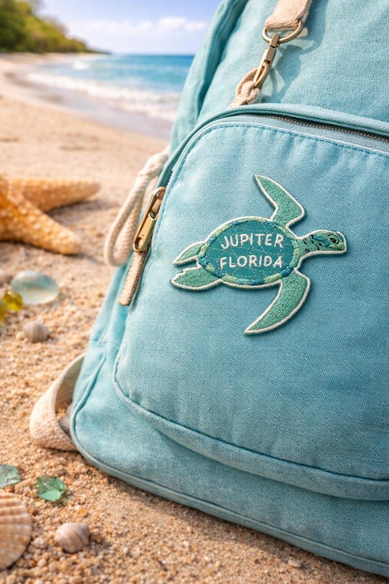 Pu&ograve; includere: Zaino azzurro con una toppa a forma di tartaruga con la scritta "Jupiter Florida". Lo zaino ha una tasca frontale con cerniera dorata e spallacci beige. Lo sfondo mostra una spiaggia sabbiosa e l'oceano.