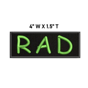 May include: A rectangular embroidered patch with a black background and the word RAD stitched in bright lime green thread. The patch features a black border and includes the printed text 4" W X 1.5" T above the design to indicate its dimensions.