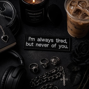 May include: A dark, gothic-inspired arrangement with a lit candle, iced coffee, and accessories. A patch reads "I'm always tired, but never of you." Also visible are rings, a necklace, and a black rose.
