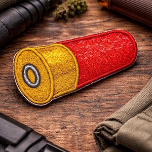 Può includere: Toppa ricamata di un bossolo di fucile, con base gialla e corpo rosso. La toppa è bordata con cuciture gialle e poggia su una superficie di legno, con armi da fuoco e tessuto sullo sfondo.