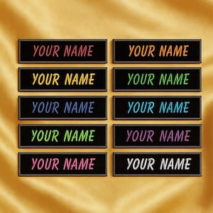 Può includere: Dieci toppe ricamate con la scritta "YOUR NAME" in vari colori, tra cui rosso, arancione, verde e blu, su sfondo nero. Le toppe sono disposte su un tessuto dorato.