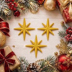 Puede incluir: Tres adornos en forma de estrella de ocho puntas de color dorado con una apariencia texturizada y cosida. Cada estrella presenta un punto central con rayos radiantes, dispuestos en formación triangular sobre una superficie de madera clara.