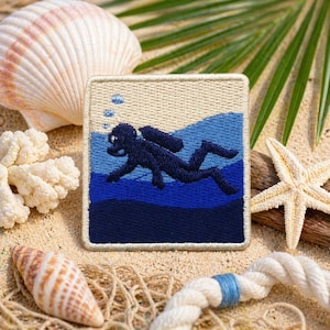 May include: Embroidered patch featuring a diver in dark blue against a blue and beige background. The patch is square with a stitched border. The image is set on a sandy surface with seashells, coral, and a starfish.