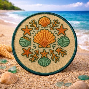 May include: A round embroidered patch featuring a seashell design in shades of orange, green, and beige. The patch has a dark green border and includes starfish and coral elements. The background shows a beach scene with sand and ocean.