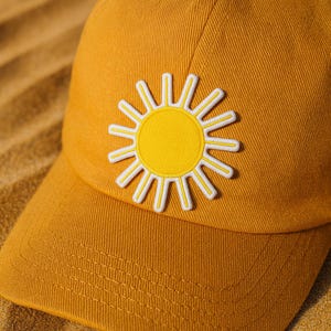 May include: Mustard yellow baseball cap with a bright yellow sun patch with white outline. The cap has a curved brim and a structured crown. The sun patch is centered on the front of the cap, adding a cheerful touch to the accessory.
