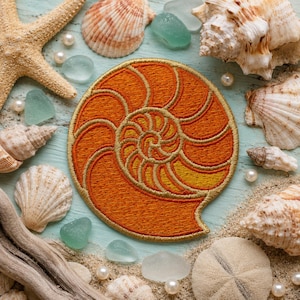 Peut inclure: Un écusson brodé circulaire représentant un motif de coquille de nautile dans des tons orange et jaune avec des contours en fil doré. L'écusson est entouré de divers coquillages, de perles blanches et de morceaux de verre de mer polis.