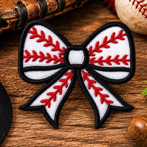 Puede incluir: Un lazo con temática de béisbol con un centro blanco, costuras rojas y un contorno negro. El lazo está diseñado para parecerse a una pelota de béisbol. La imagen también incluye una pelota de béisbol, un guante de béisbol y un bate de béisbol de madera.