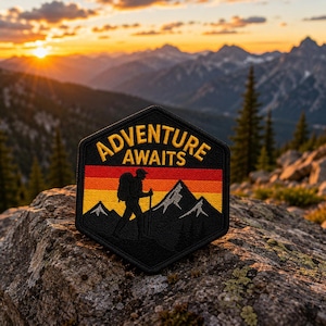 Może przedstawiać: Czarna, sześciokątna naszywka z żółtym napisem "ADVENTURE AWAITS". Naszywka przedstawia sylwetkę turysty z plecakiem i kijem, na tle gór i zachodu słońca.