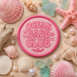 May include: A circular pink embroidered patch featuring the text OCEAN BABE CLUB in a wavy font, accented with two small hearts and wavy lines. The patch is surrounded by various seashells, a pink starfish, and white pearls.