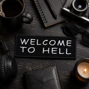 Puede incluir: Un parche rectangular negro bordado con el texto blanco WELCOME TO HELL en una fuente sans-serif en negrita. El parche tiene un borde cosido limpio y está colocado centralmente entre varios objetos personales y accesorios oscuros.