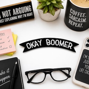 May include: A black fabric patch shaped like a banner with the white embroidered text OKAY BOOMER. The patch features a curved design with pointed ends, showcasing clean white stitching against a solid black background, suitable for attachment.