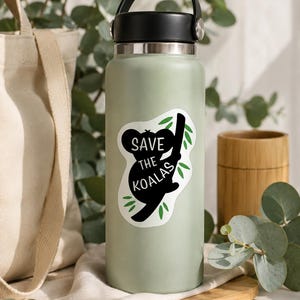 May include: A light green water bottle with a black handle and lid. A white sticker with a black koala silhouette and the text "Save the Koalas" is affixed to the bottle. Green leaves accent the koala and text.