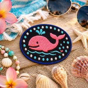 May include: An oval patch featuring a pink whale design on a black background, surrounded by white dots and a blue border. The patch is set on a wooden surface with beach-themed accessories like sunglasses, seashells, and a starfish.
