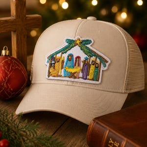 May include: A beige trucker cap featuring a central embroidered patch of a nativity scene. The patch depicts figures in blue, yellow, purple, and green robes under a green roof structure with a yellow star at the peak, all on a light fabric cap.