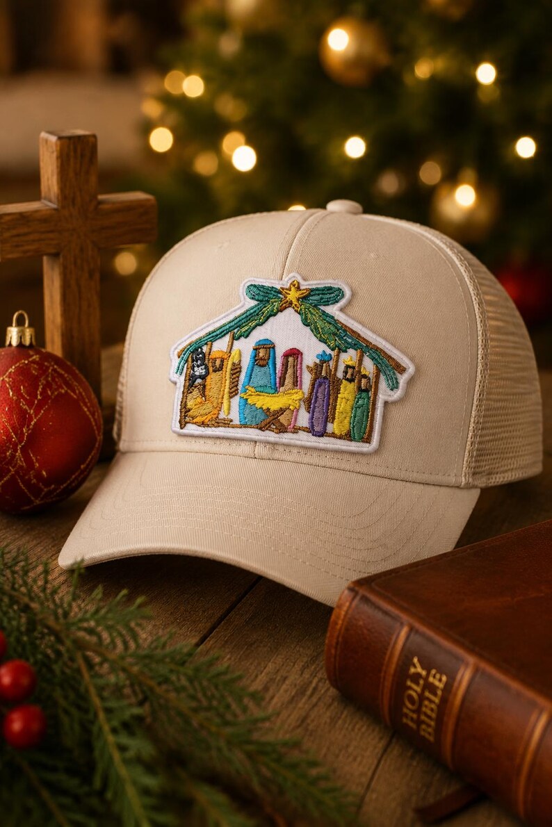 May include: A beige trucker cap featuring a central embroidered patch of a nativity scene. The patch depicts figures in blue, yellow, purple, and green robes under a green roof structure with a yellow star at the peak, all on a light fabric cap.