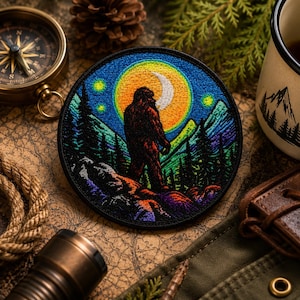 May include: Round embroidered patch featuring a Bigfoot silhouette against a night sky with a crescent moon and stars. The design includes mountains and pine trees in shades of green, blue, and purple. The patch has a black border.