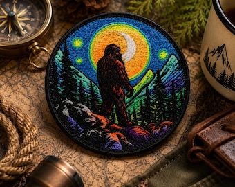 May include: Round embroidered patch featuring a Bigfoot silhouette against a night sky with a crescent moon and stars. The design includes mountains and pine trees in shades of green, blue, and purple. The patch has a black border.