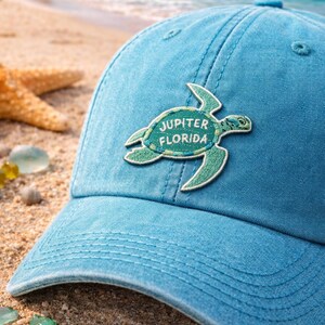 Pu&ograve; includere: Un cappellino da baseball azzurro con una toppa a forma di tartaruga verde con la scritta "Jupiter Florida". Il cappellino &egrave; realizzato in tessuto morbido ed &egrave; appoggiato su una spiaggia sabbiosa con conchiglie e una stella marina.