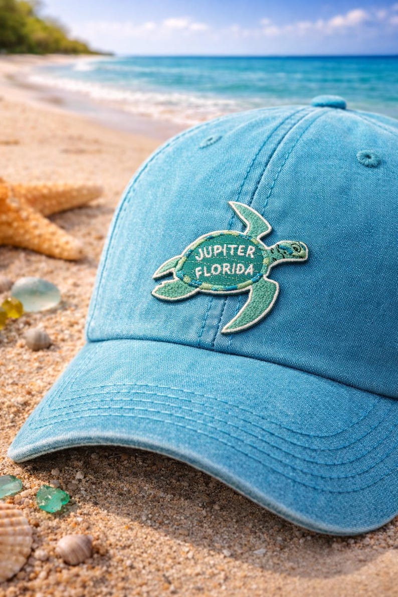 Pu&ograve; includere: Un cappellino da baseball azzurro con una toppa a forma di tartaruga verde con la scritta "Jupiter Florida". Il cappellino &egrave; realizzato in tessuto morbido ed &egrave; appoggiato su una spiaggia sabbiosa con conchiglie e una stella marina.