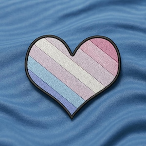 以下が含まれることがあります： 黒い縁取りの刺繍ハートパッチ。ハートは、ピンク、白、青の色合いの水平ストライプのデザインが特徴です。パッチは青色の布の背景に配置されています。