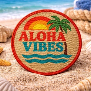 May include: Round patch with the words "ALOHA VIBES" in red and teal, featuring a palm tree, waves, and a sunset design. The patch is on a sandy surface with seashells and a starfish.