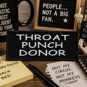 May include: A rectangular black embroidered patch with white text that reads THROAT PUNCH DONOR. The patch is surrounded by various desk items including a black mug, a yellow smiley face, a skeleton figurine, and several notepads with text.
