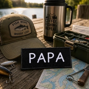 May include: An olive green baseball cap with a patch reading FISH MORE WORRY LESS, a silver travel mug with COFFEE FISHING REPEAT text, a black rectangular patch with white PAPA text, a green tackle box, fishing lures, and a map.