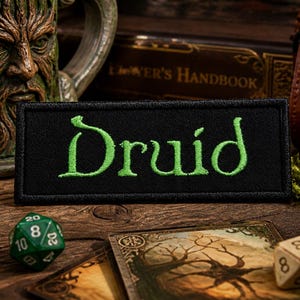 Könnte beinhalten: Ein schwarzer Aufnäher mit dem Wort "Druid" in Grün gestickt, auf einer Holzoberfläche mit Würfeln, Karten und einem Becher. Der Aufnäher ist rechteckig und die Schrift ist in einer stilisierten Schriftart gehalten. Die Szene erinnert an ein Fantasy-Thema.