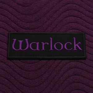 K&ouml;nnte beinhalten: Ein rechteckiger schwarzer Aufn&auml;her mit dem Wort Warlock, das in violettem Garn gestickt ist. Der Aufn&auml;her ist auf einem tiefvioletten Stoffhintergrund zentriert, der eine texturierte, wellenf&ouml;rmige und erhabene Struktur aufweist.